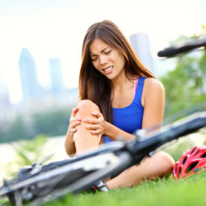 Knee pain bike injury. Woman with pain in knee joints after biking on bicycle. Girl sitting down with painful face expression. Mixed race sport fitness model outdoors.